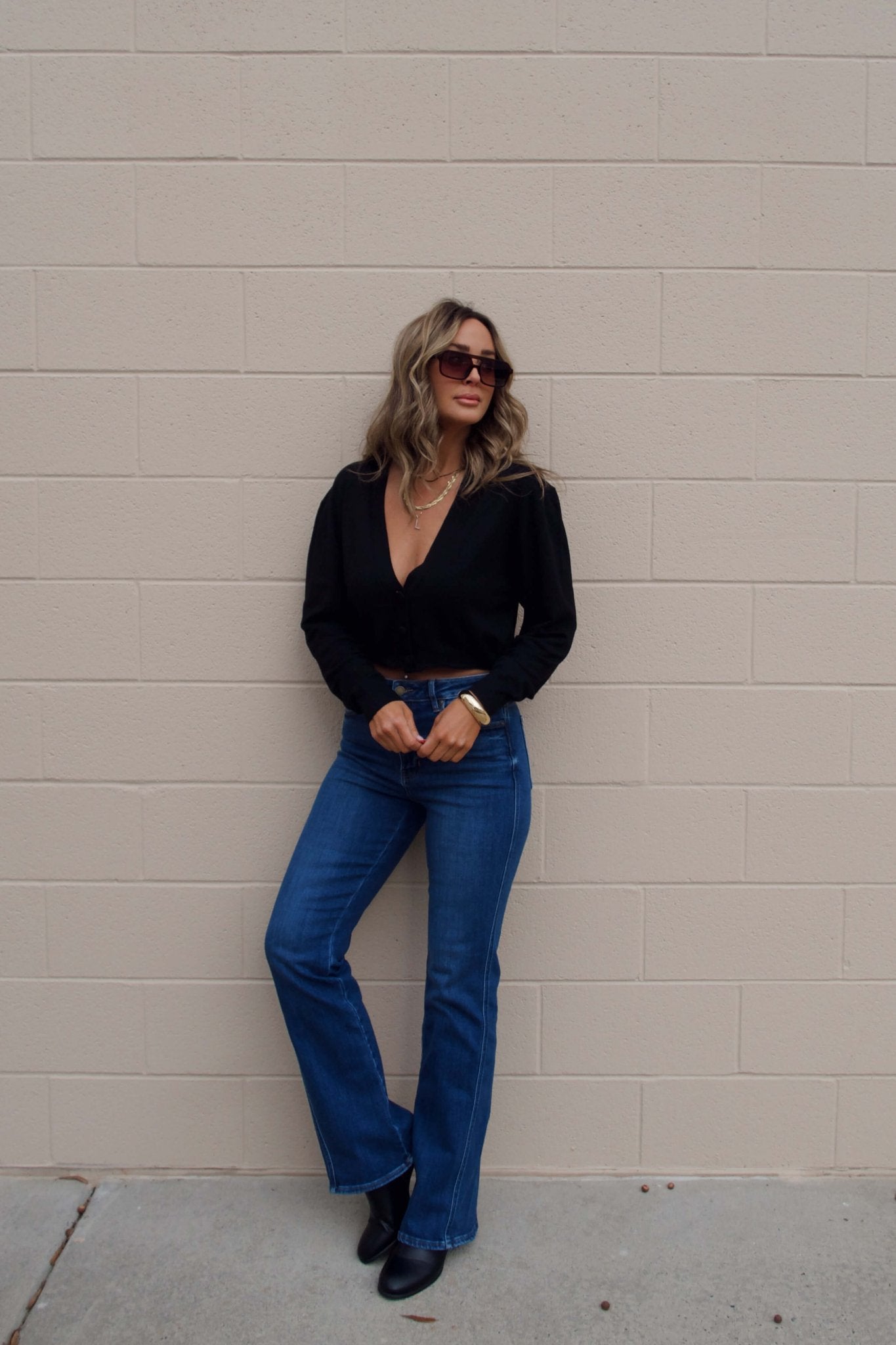 Woman with wavy hair in sunglasses and blue jeans models the Black V Neck Buttoned Cardigan against a beige brick wall.