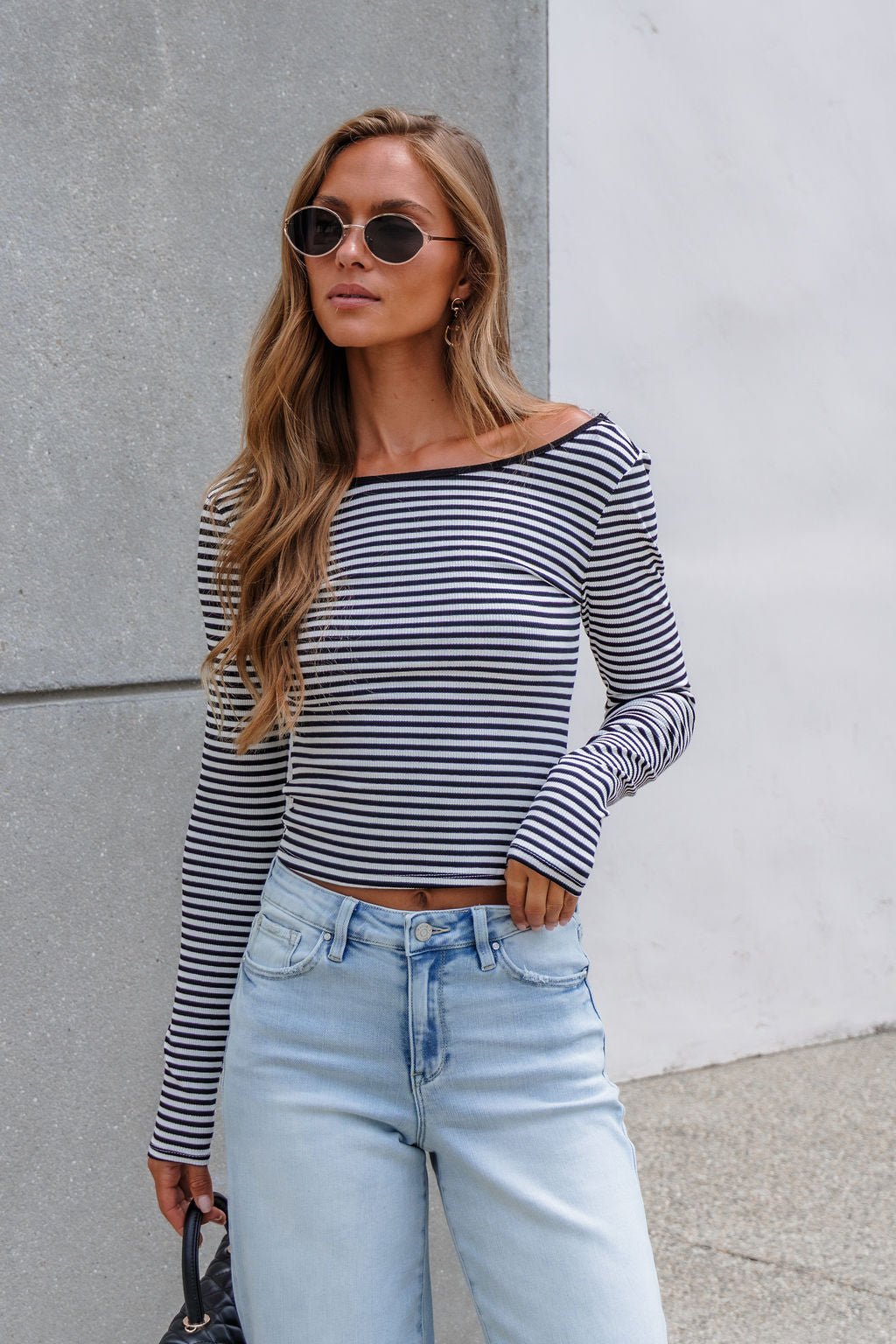 Woman with long hair in sunglasses wears a Black & White Striped Boatneck Top with light blue jeans by a concrete and white wall.