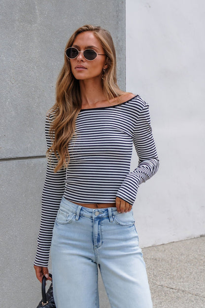 Woman with long hair in sunglasses wears a Black & White Striped Boatneck Top with light blue jeans by a concrete and white wall.