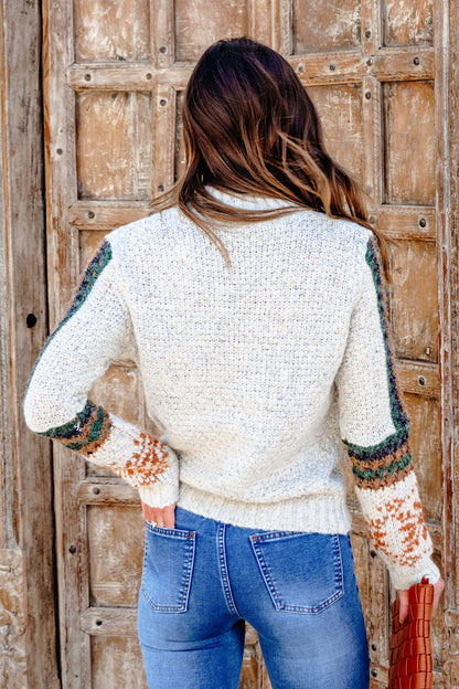 Woman with long brown hair in the Blaire Oatmeal Fair Isle Sweater and blue jeans stands by a rustic wooden door, embracing fall style.