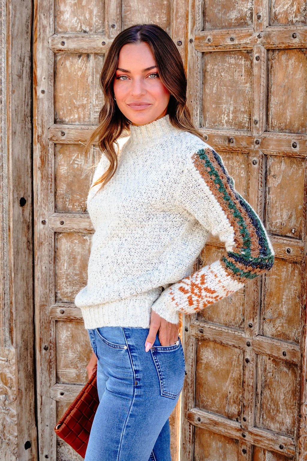Woman in a Blaire Oatmeal Fair Isle Sweater and blue jeans stands by a rustic door with a purse—perfect for fall style.