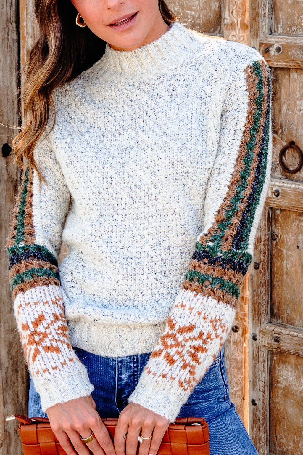 Woman in the Blaire Oatmeal Fair Isle Sweater, holding a brown clutch by a wooden door—perfect for fall style.
