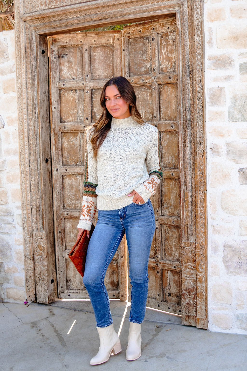 Woman in the Blaire Oatmeal Fair Isle Sweater, blue jeans, and boots stands by a rustic door holding a brown clutch.