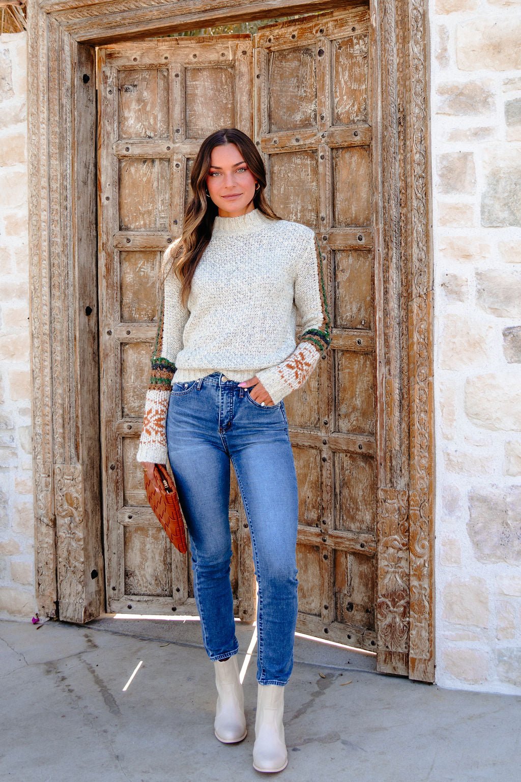 Woman in the Blaire Oatmeal Fair Isle Sweater, blue jeans, and white boots holds a brown clutch by a rustic wooden door.