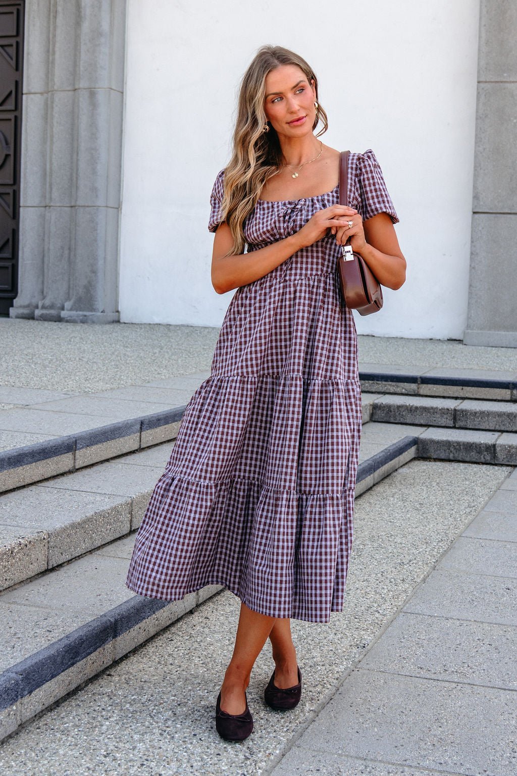 Woman wears BLU PEPPER B6FD1108 MOCHA MIDI DRESS on steps, holding a brown handbag, with long wavy hair and a neutral expression.