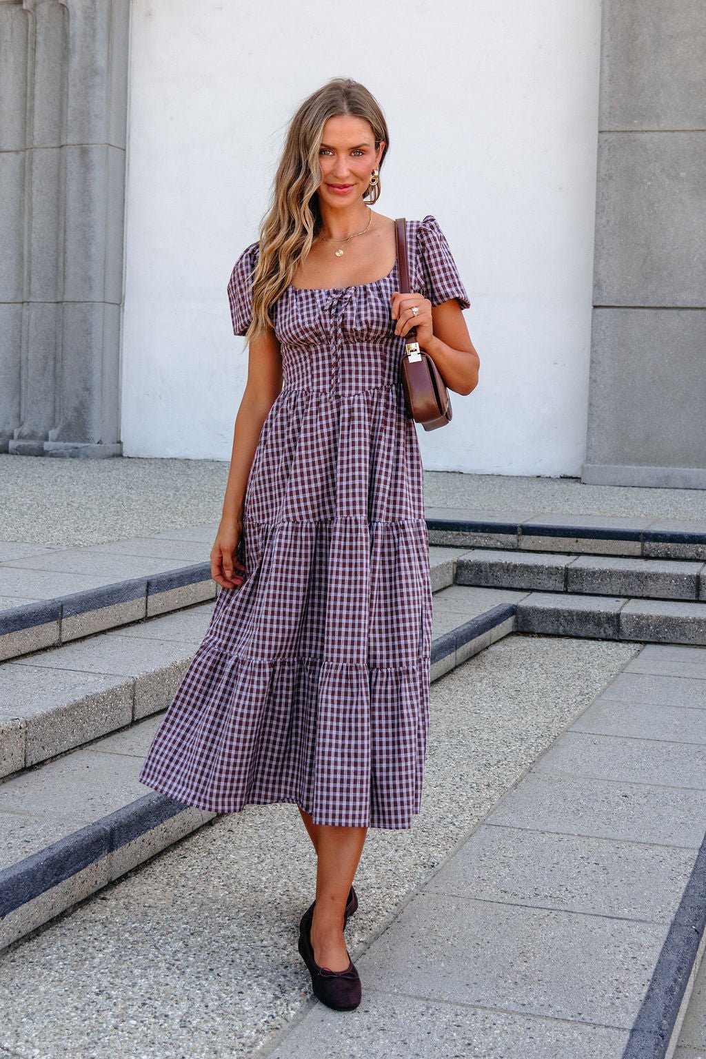 Woman in a BLU PEPPER B6FD1108 MOCHA MIDI DRESS stands on steps, holding a brown purse and smiling at the camera.