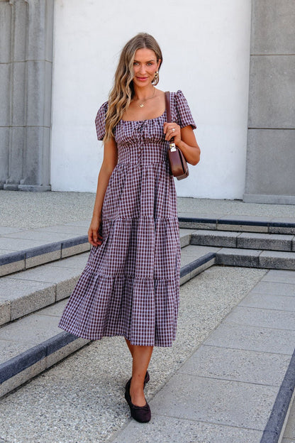 Woman in a BLU PEPPER B6FD1108 MOCHA MIDI DRESS stands on steps, holding a brown purse and smiling at the camera.