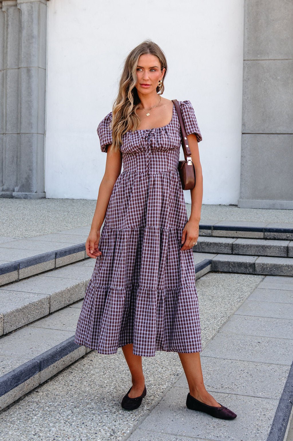 Woman in a BLU PEPPER B6FD1108 MOCHA MIDI DRESS stands on stone steps outdoors, holding a brown bag and wearing black flats.