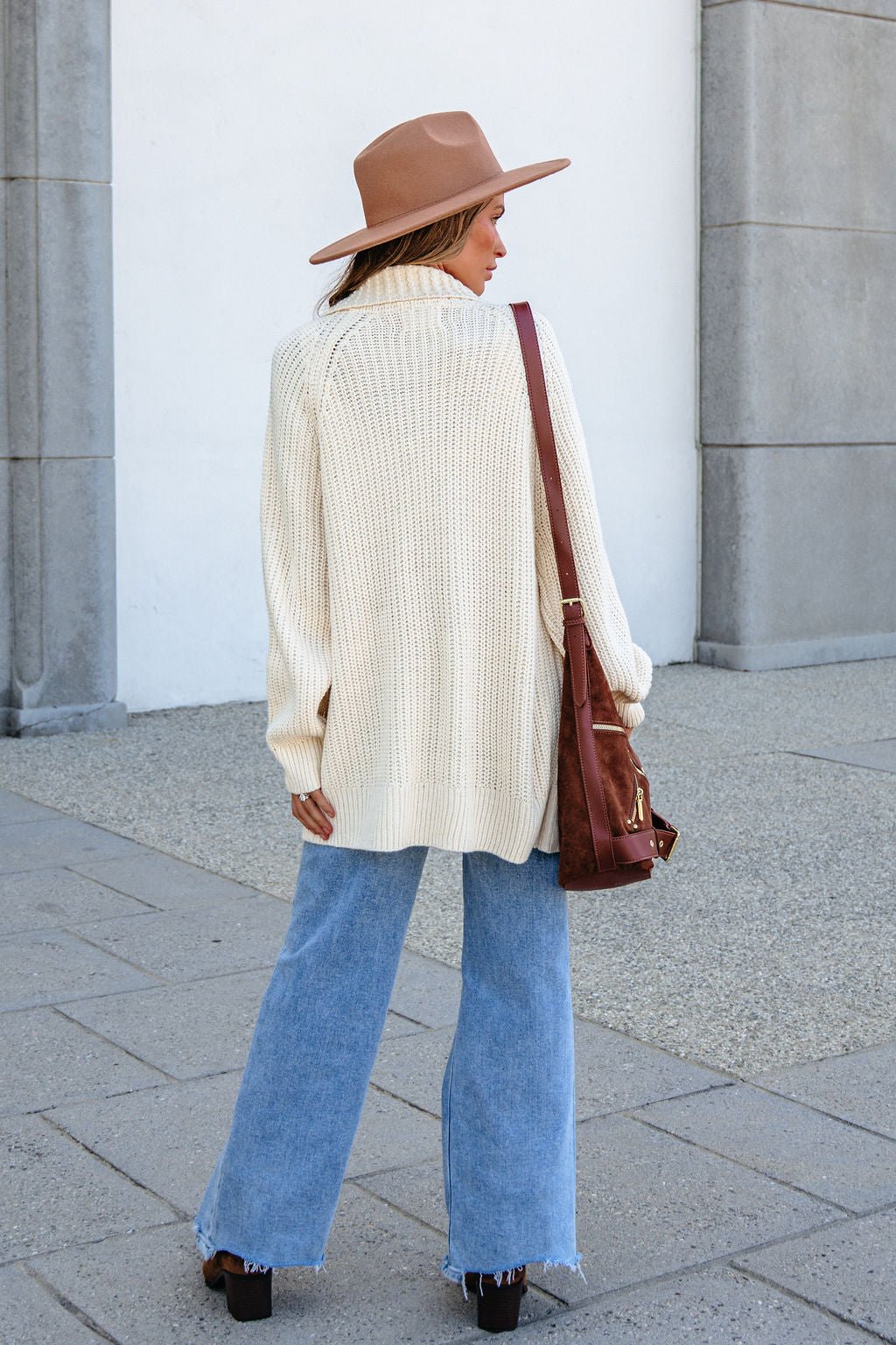 Woman in a beige hat, BLU PEPPER J6FJ1011 IVORY SWEATER, and light blue jeans stands outdoors with a brown bag.