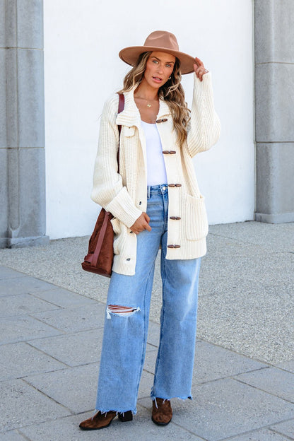 Woman in BLU PEPPER J6FJ1011 IVORY SWEATER, tan hat, ripped jeans, brown boots, and bag stands outdoors by a gray wall.