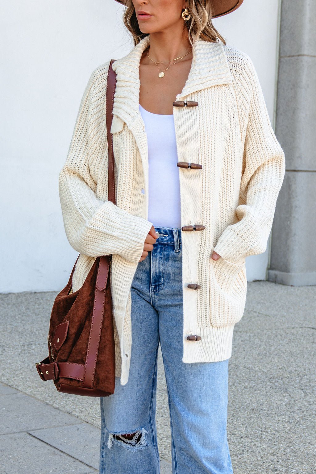 Woman wearing BLU PEPPER J6FJ1011 IVORY SWEATER, white top, ripped jeans, brown hat, and carrying a brown shoulder bag.