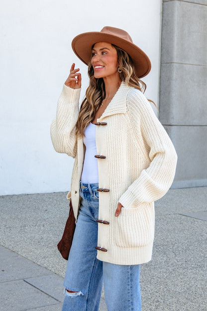 Woman in BLU PEPPER J6FJ1011 IVORY SWEATER, blue jeans, and hat smiles while standing outside by a light wall.