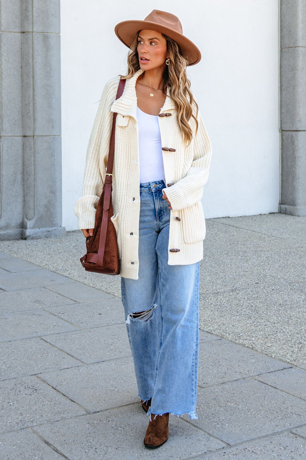Woman in a tan hat, BLU PEPPER J6FJ1011 IVORY SWEATER, white top, ripped jeans, brown boots, and brown bag walking outdoors.