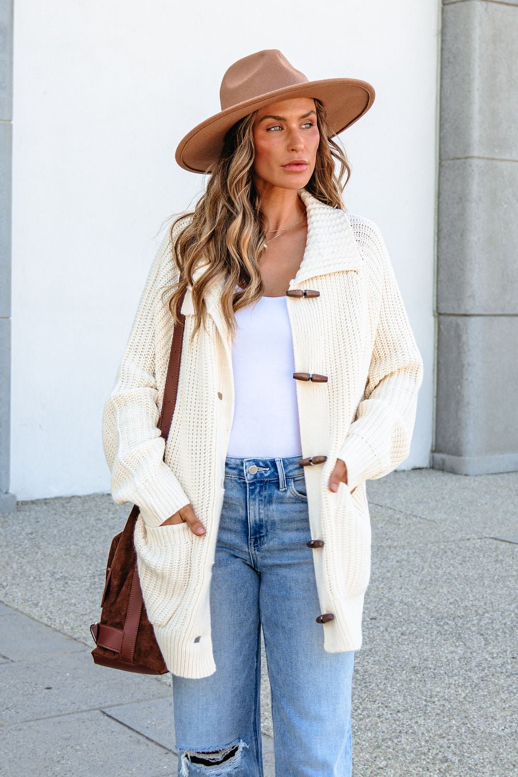 Woman wears BLU PEPPER J6FJ1011 IVORY SWEATER, tan hat, white top, jeans, and carries a brown bag outdoors with hands in pockets.