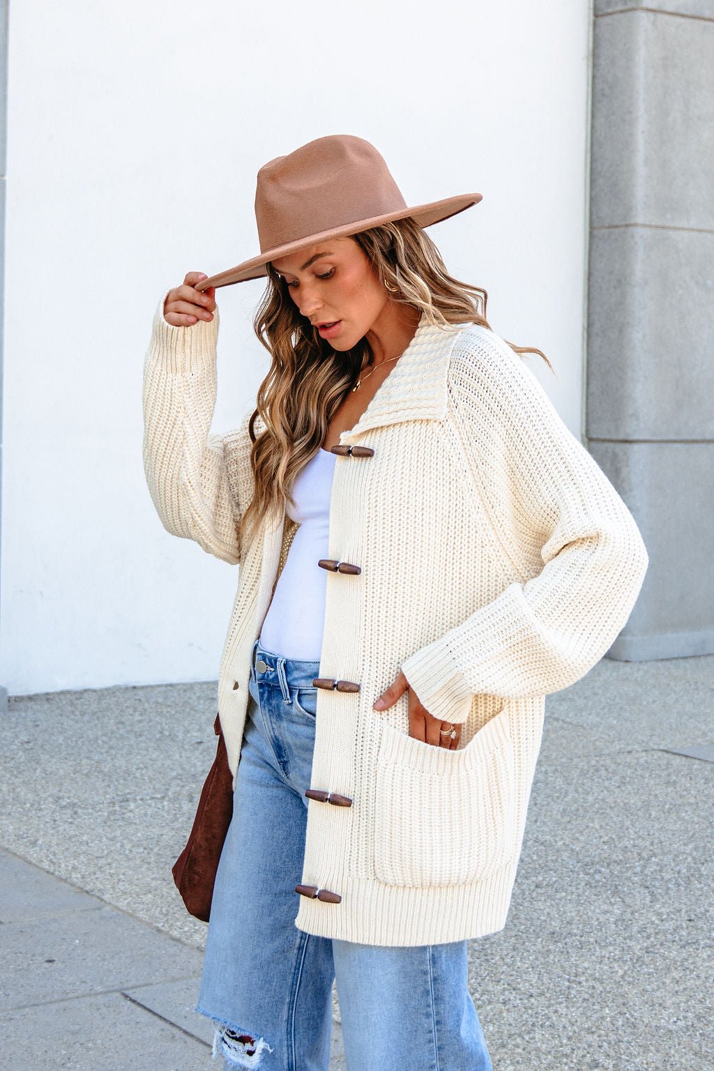 Woman wears BLU PEPPER J6FJ1011 IVORY SWEATER, ripped jeans, and tan hat outdoors, looking down with hand on her hat.