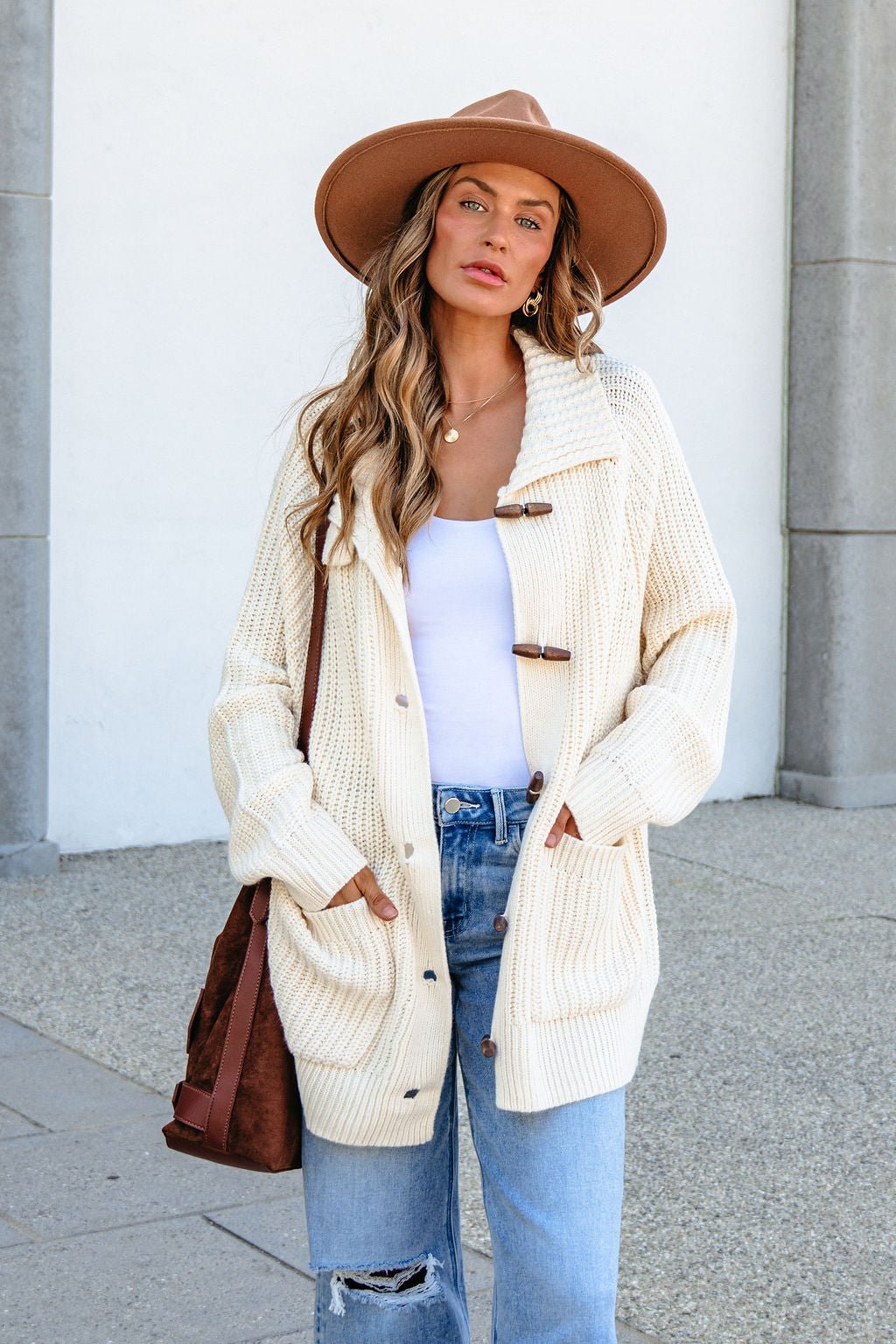 Woman in BLU PEPPER J6FJ1011 IVORY SWEATER, white top, jeans, tan hat, with a brown bag stands outside with hands in pockets.