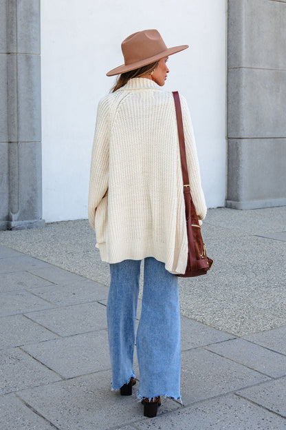Woman in a wide-brim hat, BLU PEPPER J6FJ1011 IVORY SWEATER, flared jeans, black shoes, and brown shoulder bag stands outside.