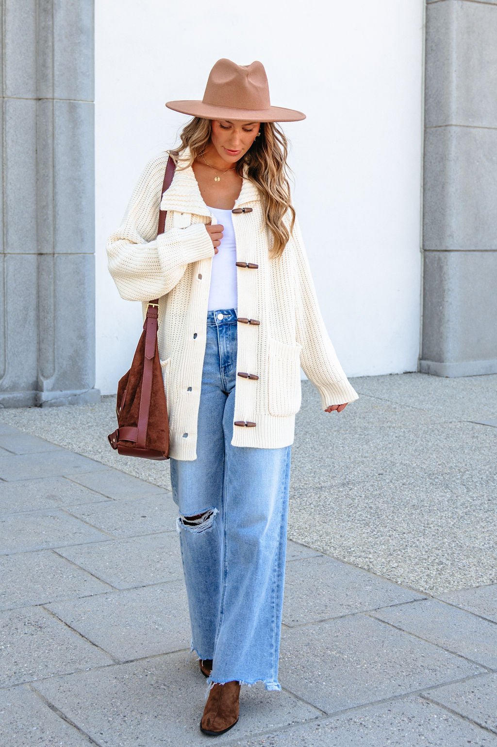 Woman in a tan hat, BLU PEPPER J6FJ1011 IVORY SWEATER, white top, ripped jeans, brown boots, and brown shoulder bag walks outside.