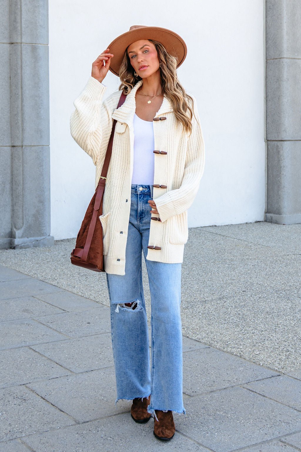 Woman in wide-brimmed hat, BLU PEPPER J6FJ1011 IVORY SWEATER, white top, ripped jeans, boots, and crossbody bag by stone columns.