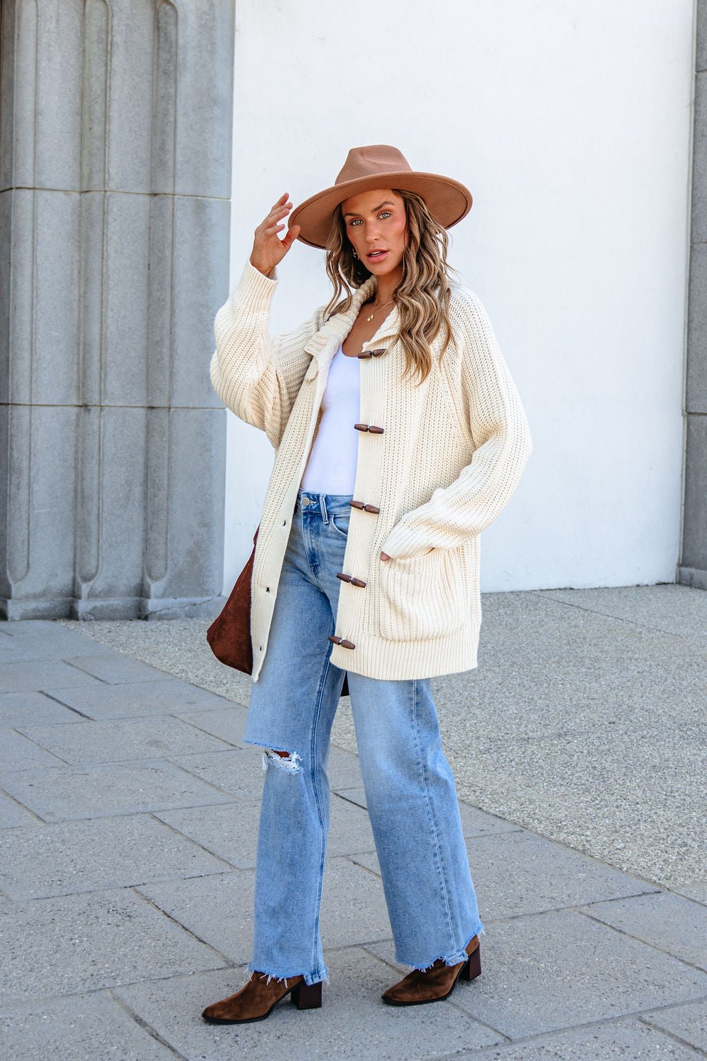 Woman in BLU PEPPER J6FJ1011 IVORY SWEATER, jeans, tan hat, and boots stands by stone columns holding her hat.