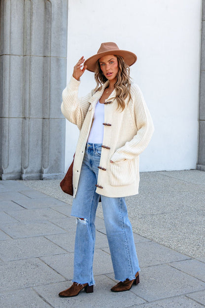 Woman in BLU PEPPER J6FJ1011 IVORY SWEATER, jeans, tan hat, and boots stands by stone columns holding her hat.