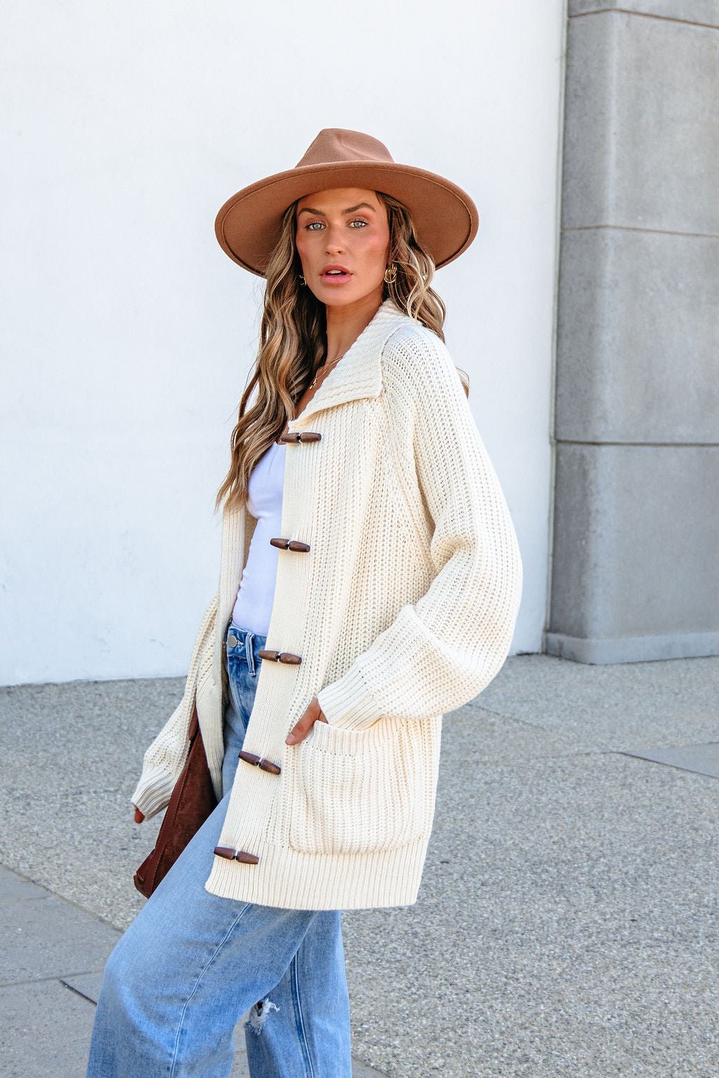 Woman in tan hat, blue jeans, and a BLU PEPPER J6FJ1011 IVORY SWEATER stands outdoors against a light wall, facing the camera.