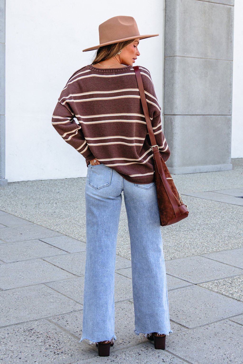Woman in BLU PEPPER J6FT1014 brown striped sweater, light blue jeans, tan hat, and brown bag, standing outdoors facing away.