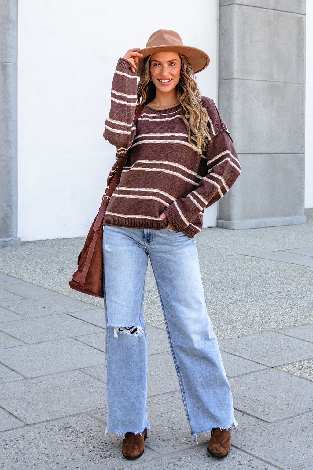 Woman in the BLU PEPPER J6FT1014 Brown Striped Sweater stands outside, smiling, with a brown bag and tan hat.