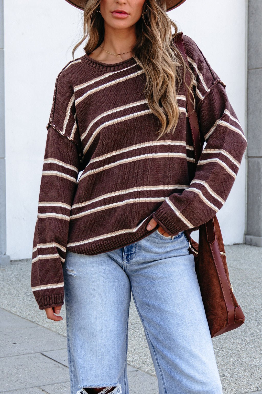 Woman in a BLU PEPPER J6FT1014 brown striped sweater, light blue ripped jeans, and brown hat outdoors, holding a matching brown bag.