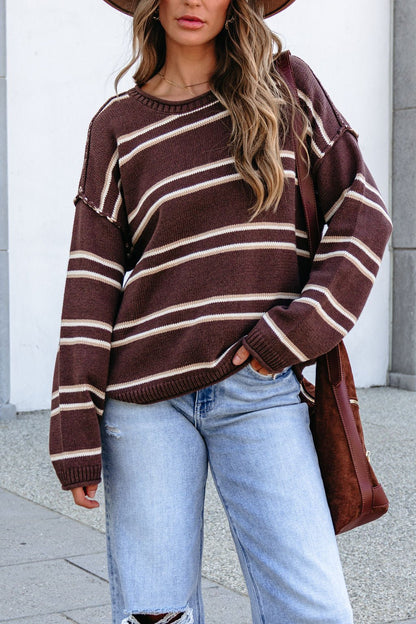 Woman in a BLU PEPPER J6FT1014 brown striped sweater, light blue ripped jeans, and brown hat outdoors, holding a matching brown bag.