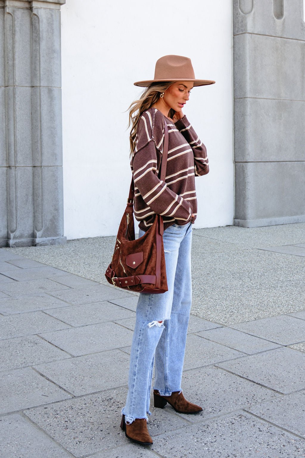 Woman in the BLU PEPPER J6FT1014 brown striped sweater, brown hat, ripped jeans, and boots stands by a stone building with a brown bag.
