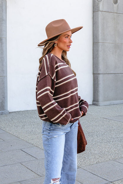 Woman wears BLU PEPPER J6FT1014 brown striped sweater, tan hat, and ripped jeans outside a gray and white building.