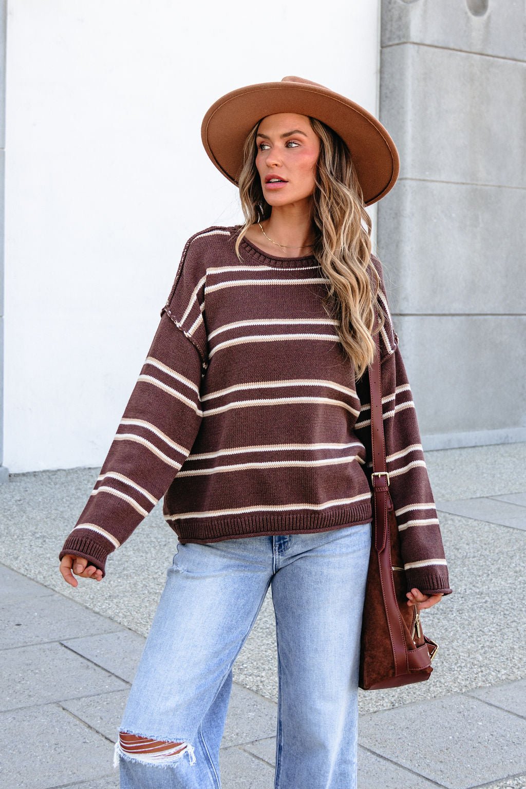 Woman in BLU PEPPER J6FT1014 brown striped sweater, hat, ripped jeans, carrying a brown shoulder bag outdoors.