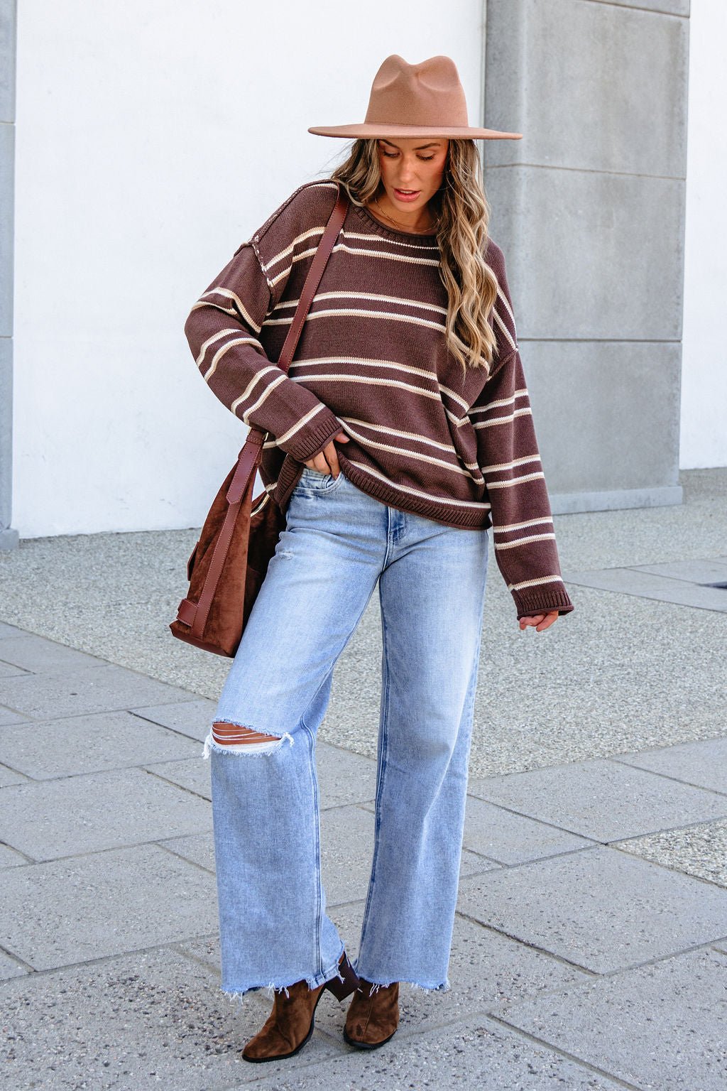 Woman wears BLU PEPPER J6FT1014 BROWN STRIPED SWEATER, jeans, brown hat, and boots, holding a brown bag on the sidewalk.