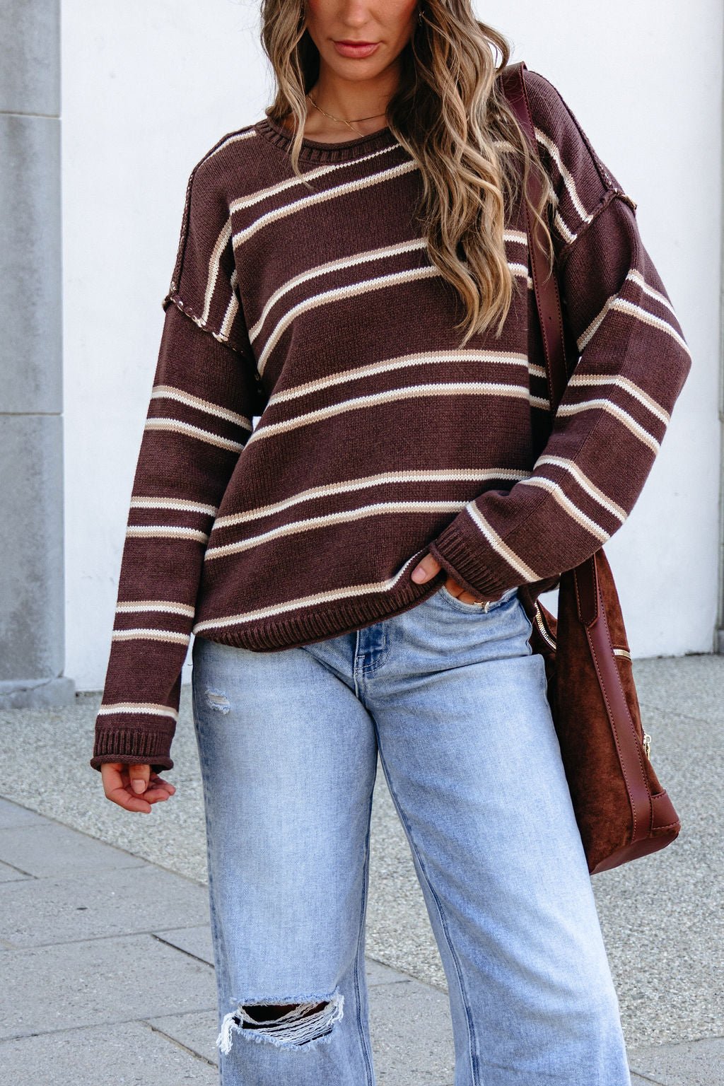 Woman in a BLU PEPPER J6FT1014 brown striped sweater and ripped jeans stands outdoors, holding a brown bag.