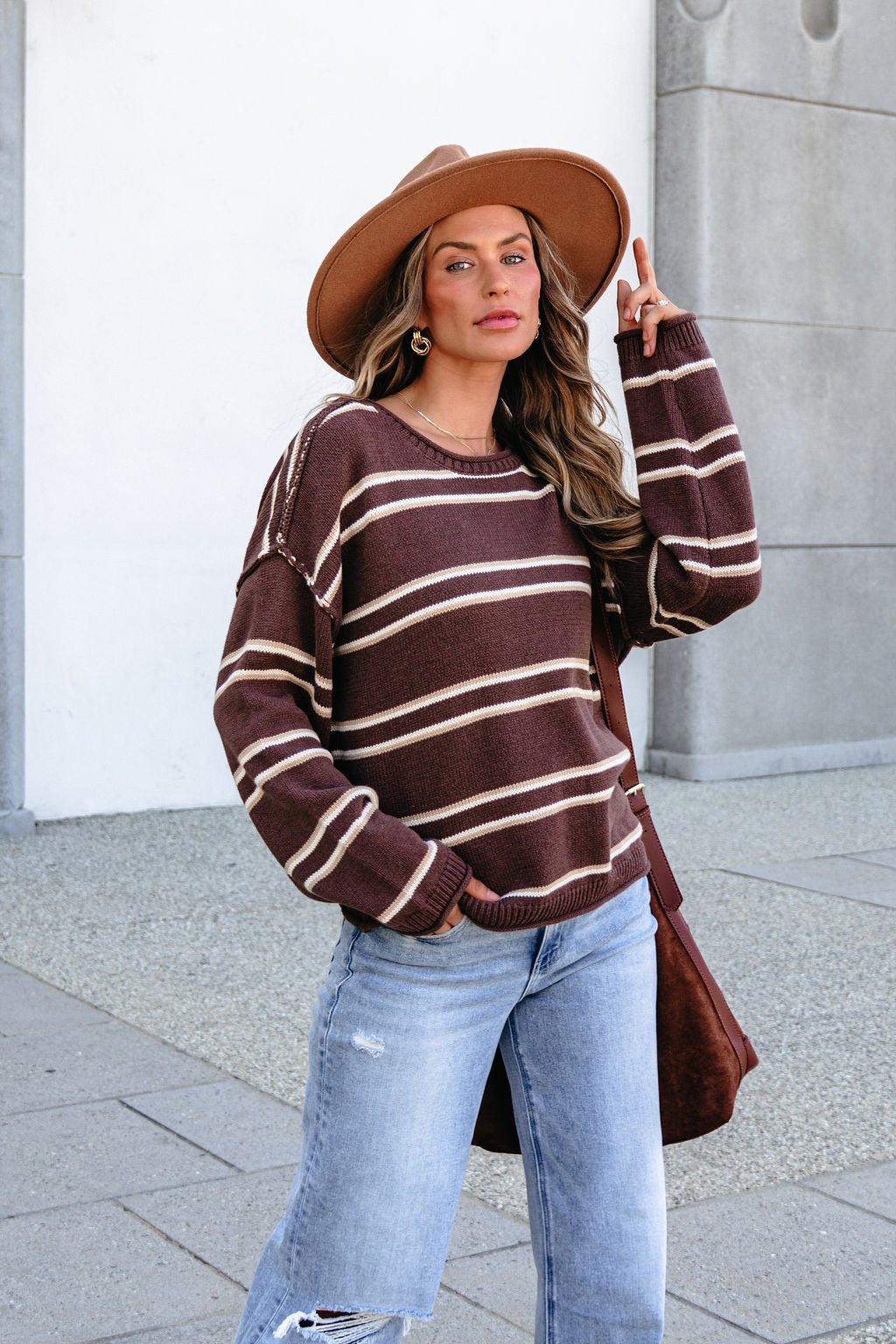 Woman wears BLU PEPPER J6FT1014 brown striped sweater, ripped jeans, & a hat outdoors, holding her hat and looking confidently at the camera.