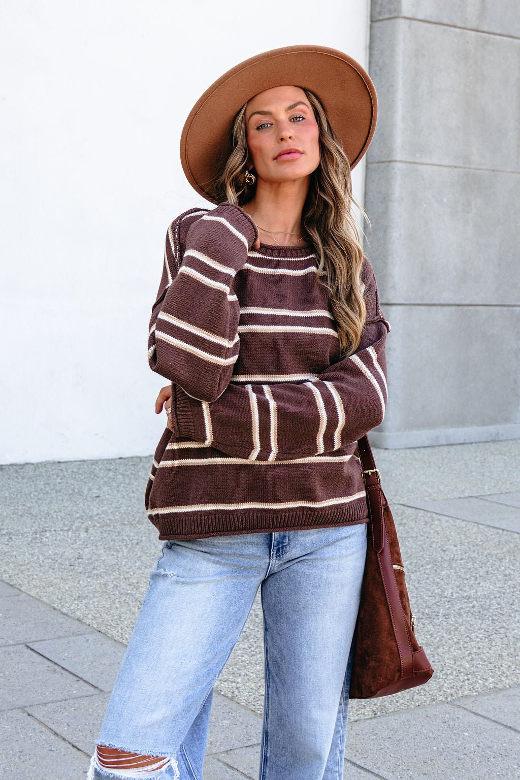 Woman in BLU PEPPER J6FT1014 brown striped sweater, light blue ripped jeans, tan hat, and brown shoulder bag stands outside.