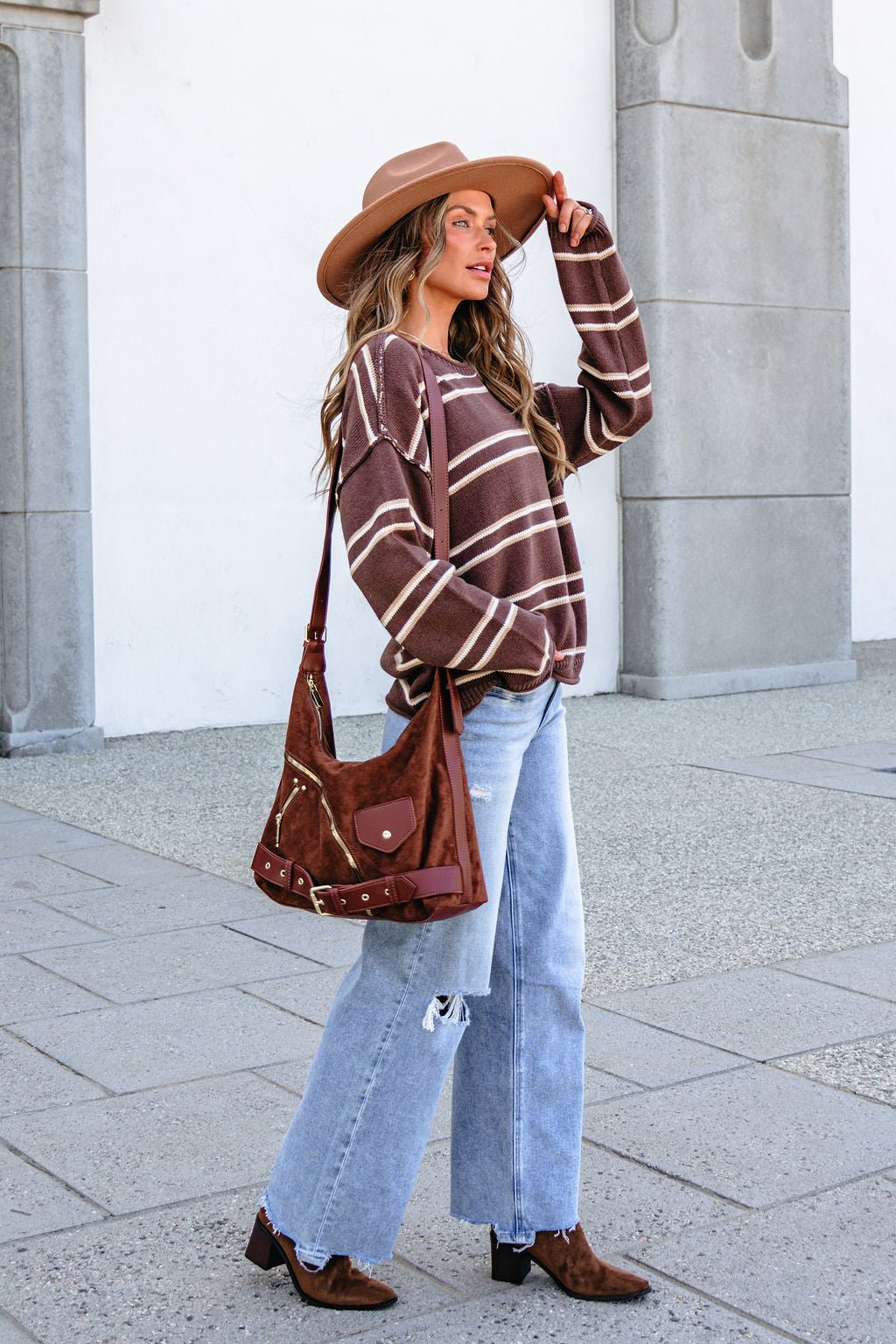 Woman in BLU PEPPER J6FT1014 brown striped sweater stands outside with ripped jeans, boots, wide-brim hat, and a brown shoulder bag.