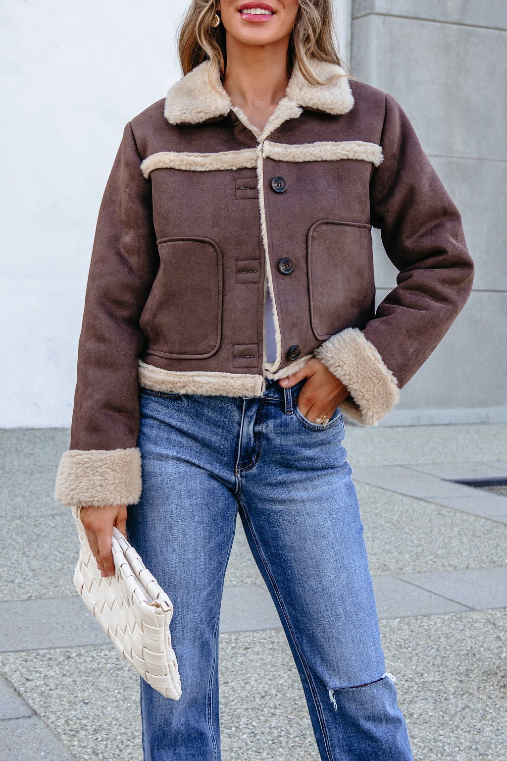 Woman in BLU PEPPER TW2302 brown jacket and blue jeans holds a white woven clutch while standing outdoors.