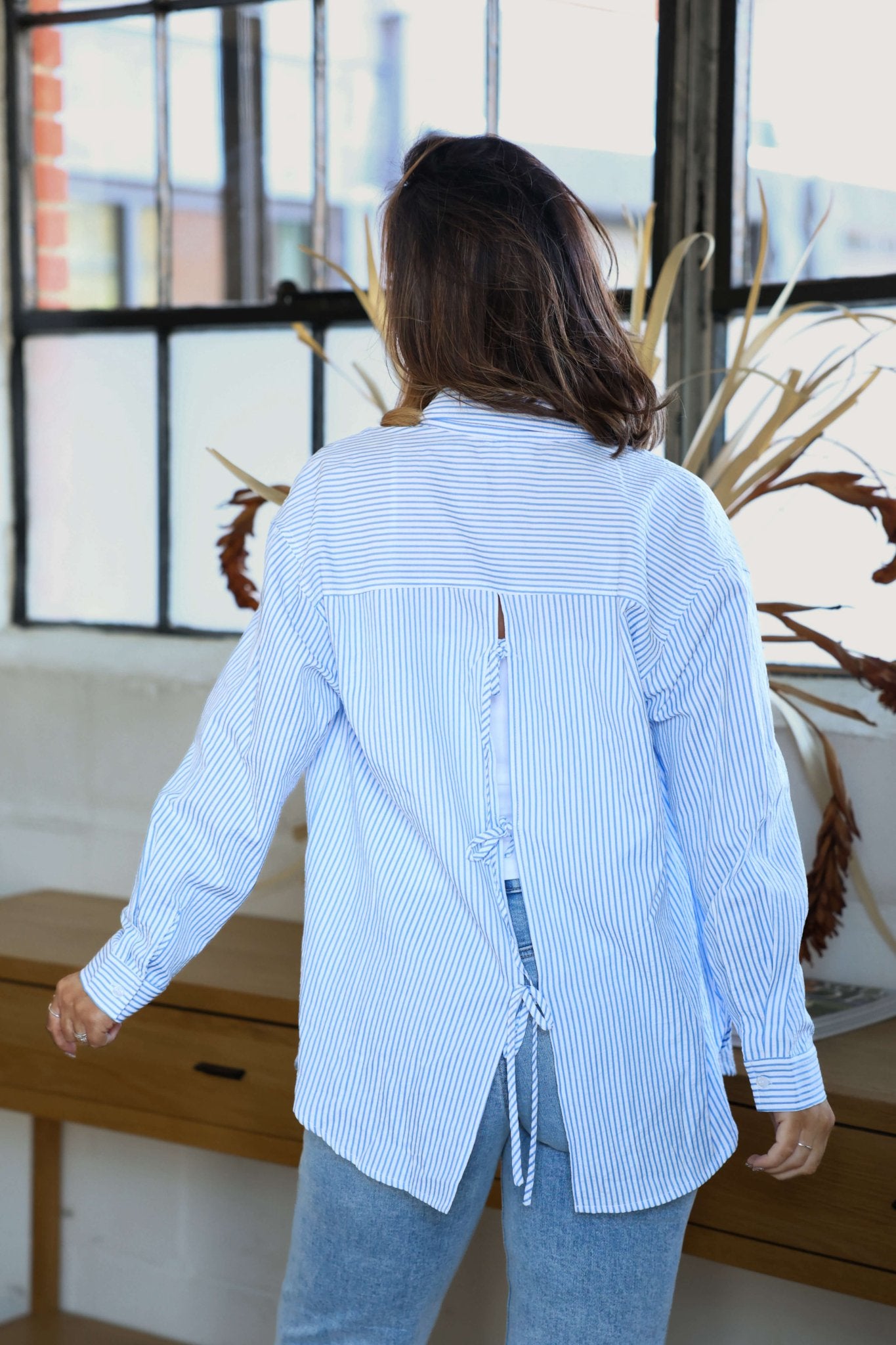 A woman wears the Blue and White Striped Tie Top, standing with her back to the camera in a bright room with large windows.