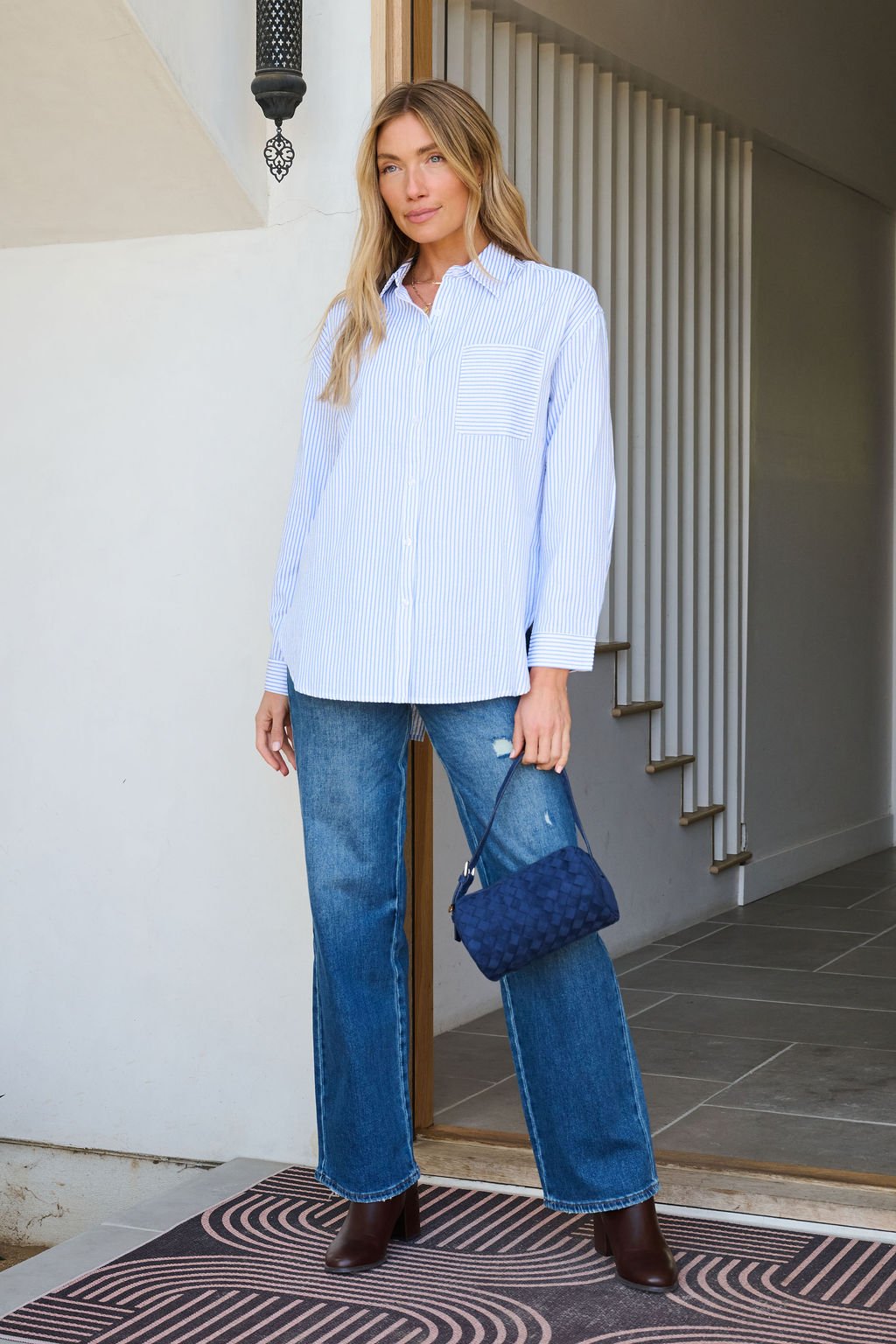 Woman stands indoors wearing the Blue and White Striped Tie Top, blue jeans, brown boots, and a blue quilted handbag.