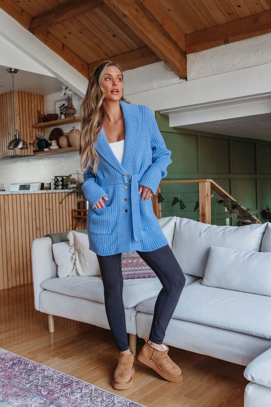 Woman wears a Blue Button Down Belted Cardigan–FINAL SALE, white top, black leggings & tan boots in a cozy modern living room.