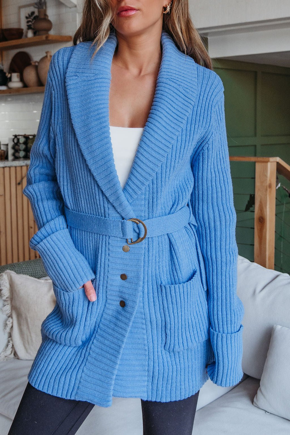 A woman wears the Blue Button Down Belted Cardigan - FINAL SALE over a white top and black pants in a modern living room.