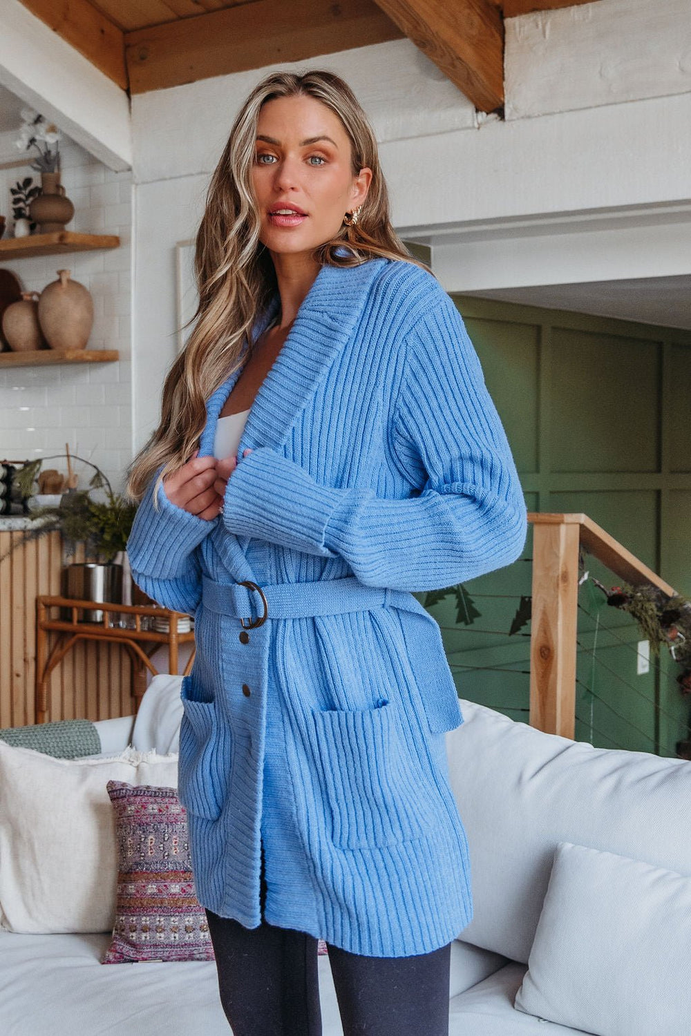 A woman models the Blue Button Down Belted Cardigan - FINAL SALE in a cozy, neutral-toned living room with wood accents.