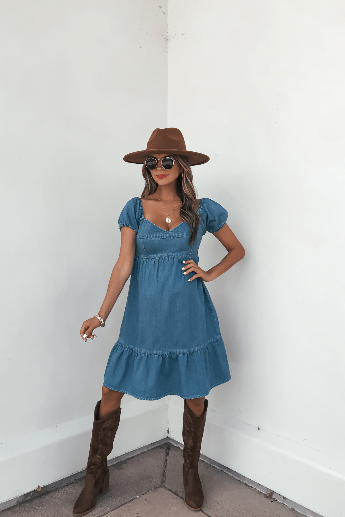 Woman in a Blue Denim Tiered Mini Dress - DOORBUSTER, brown hat, and boots stands by a white wall wearing sunglasses.