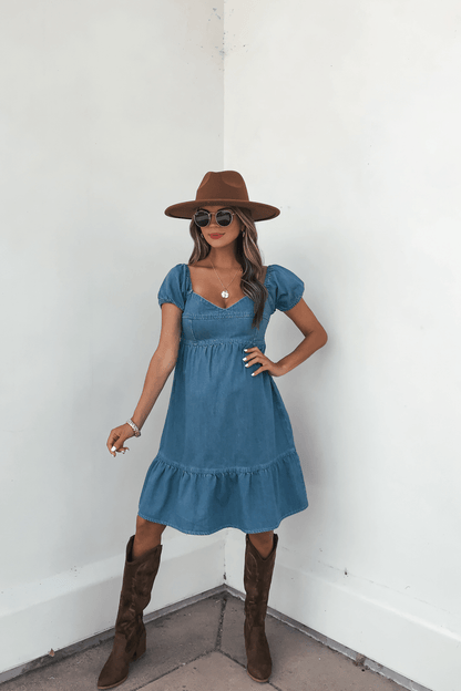 Woman in a Blue Denim Tiered Mini Dress - DOORBUSTER, brown hat, and boots stands by a white wall wearing sunglasses.