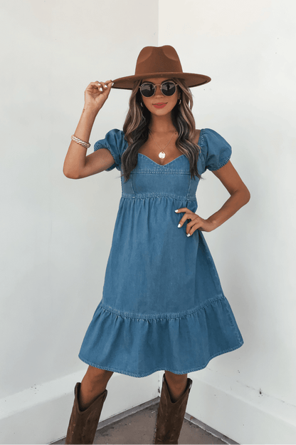 Woman in a Blue Denim Tiered Mini Dress - DOORBUSTER, brown hat, and boots poses against a light background.