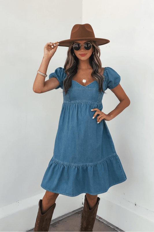 Woman in a Blue Denim Tiered Mini Dress - DOORBUSTER, brown hat, and boots poses against a light background.