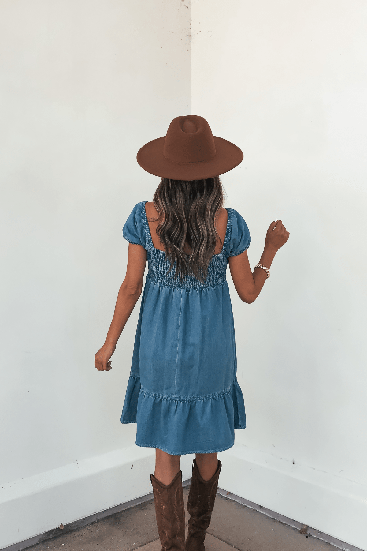 Woman with long wavy hair in a brown hat and boots wears the Blue Denim Tiered Mini Dress - DOORBUSTER, facing a white wall.