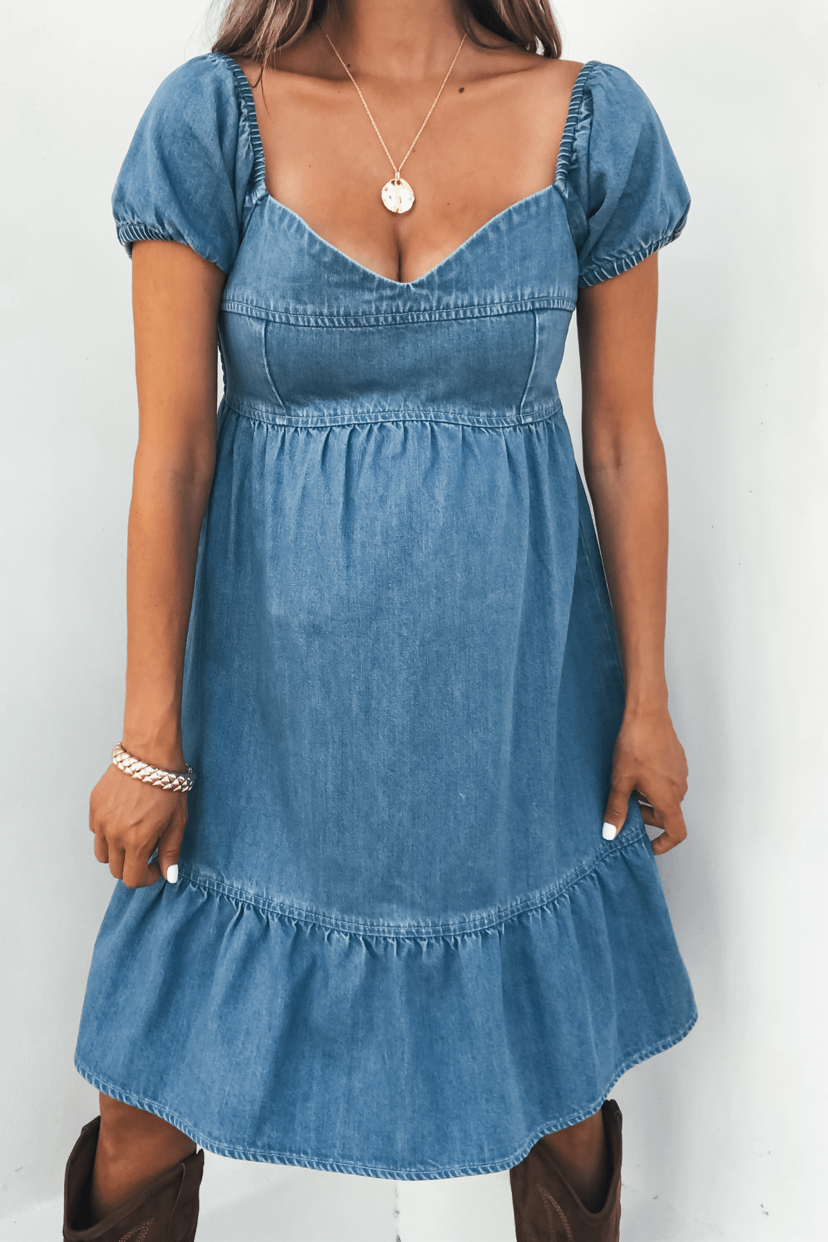 Woman in Blue Denim Tiered Mini Dress - DOORBUSTER, with a pendant necklace, bracelet, and brown boots against a plain background.
